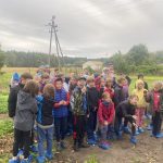 Grupa dzieci w niebieskich ochraniaczach na buty stoi na zewnątrz w pochmurny dzień, otoczona zielenią.