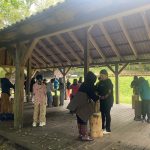 Dzieci stoją pod drewnianą wiatą, uczestnicząc w zajęciach grupowych z drewnianymi klockami na świeżym powietrzu.