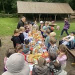 Dzieci zebrały się wokół stołu piknikowego z jedzeniem, otoczonego zielenią i drewnianą wiatą.