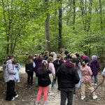 Grupa dzieci i dorosłych stoi na leśnej ścieżce otoczonej drzewami i zielenią.