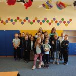 Grupa dzieci i dwoje dorosłych pozuje i uśmiecha się w kolorowej klasie z grafiką ścienną i niebieskimi szafkami.
