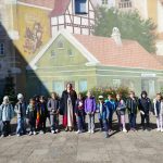 Grupa dzieci i dorosły stoją przed kolorowym muralem w słoneczny dzień.