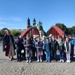 Grupa dzieci i kobieta stoją na czerwonym moście z kościołem w tle w słoneczny dzień.