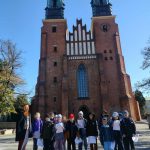 Grupa dzieci i dorosły stoją przed dużą ceglaną katedrą z wysokimi wieżami w słoneczny dzień.