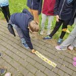 Dziecko kładzie karty z liśćmi na ziemi, podczas gdy inni stoją w kręgu na utwardzonej ścieżce.