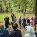 Nauczyciel rozmawia z grupą dzieci w mundurkach szkolnych podczas zajęć na świeżym powietrzu w zalesionym parku.