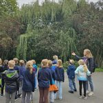 Grupa dzieci słucha kobiety przemawiającej przed drzewami w plenerze.