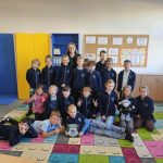 Nauczyciel i uczniowie pozują razem w klasie, uśmiechając się i siedząc na kolorowym dywanie.