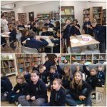 Dzieci w szkolnych mundurkach uczestniczą w zajęciach i podnoszą ręce w klasie pełnej regałów z książkami.