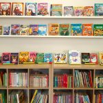 Kolorowe książki dla dzieci wyeksponowane na półkach nad dwoma regałami wypełnionymi książkami w bibliotece.