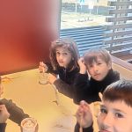 Pięcioro dzieci jedzących lody przy stoliku w restauracji, z dwójką innych dzieci siedzących przy innym stoliku w tle.