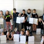 Grupa dzieci w klasie trzymających białe torby z prezentami i uśmiechających się do kamery.