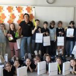 Grupa dzieci i dorosły stoją i siedzą w klasie, trzymając białe torby z prezentami i uśmiechając się do kamery.