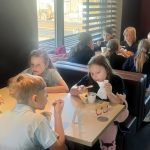 Grupa dzieci siedzi przy stolikach w restauracji typu fast food, jedząc i rozmawiając razem.