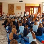 Dzieci siedzą na podłodze i obserwują grupę uczniów występujących przed szkolną salą.