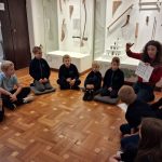 Kobieta pokazuje wykres grupie dzieci siedzących w kręgu na podłodze muzeum z instrumentami muzycznymi.