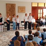 Dzieci wykonują muzykę i śpiewają przed publicznością złożoną z uczniów siedzących na podłodze w szkole.