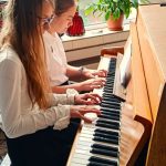 Dwie dziewczyny grające razem na pianinie przy oknie z rośliną doniczkową i światłem słonecznym wpadającym do środka.