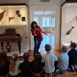 Kobieta demonstruje instrument muzyczny grupie dzieci siedzących na podłodze w eksponacie muzealnym.