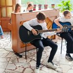 Dwoje dzieci gra na gitarach, podczas gdy inni siedzą w pobliżu; dziewczynka gra na pianinie w tle w klasie.