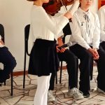 Młoda dziewczyna gra na skrzypcach, podczas gdy inni patrzą, siedząc w tle na krzesłach.