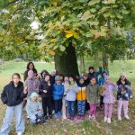 Grupa dzieci i dorosły stoją pod dużym drzewem w parku, ubrani w kolorowe jesienne ubrania.
