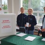 Czworo uczniów głosuje w szkolnych wyborach, wrzucając karty do urny z napisem WYBORY PRZEWODNICZĄCEGO.