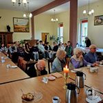 Grupa starszych osób i dzieci siedzi w jadalni ze stołami zastawionymi herbatą i ciastem.