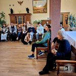 Grupa nastolatków siedzi w jasnym pokoju, podczas gdy kobieta gra na gitarze, tworząc zrelaksowaną, radosną atmosferę.