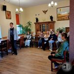 Grupa młodych ludzi siedzi i rozmawia w pokoju z drewnianymi krzesłami, dziełami sztuki i roślinami.