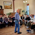 Starszy mężczyzna rozmawia z grupą siedzących nastolatków w przytulnym pokoju z roślinami i obrazem przedstawiającym krajobraz.