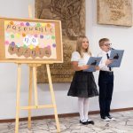 Dwoje dzieci stoi obok sztalugi z kolorowym napisem podczas szkolnej uroczystości, trzymając zeszyty.