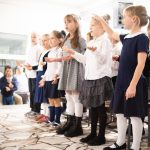 Grupa małych dzieci śpiewa i gestykuluje na scenie podczas szkolnego przedstawienia.