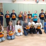 Grupa dzieci i nauczyciel trzymają udekorowane dynie w klasie i uśmiechają się do kamery.