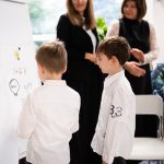 Dwóch młodych chłopców pisze na tablicy, podczas gdy dwie kobiety patrzą i uśmiechają się w jasnej klasie.