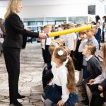 Kobieta dotyka ramienia siedzącej dziewczynki dużym, żółtym ołówkiem na oczach innych dzieci.