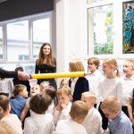 Kobieta dotyka ramienia uśmiechniętego chłopca dużym żółtym ołówkiem w klasie pełnej dzieci.