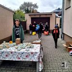 Nastoletni chłopiec stoi przy stole z zapakowanymi przedmiotami; w tle na zewnątrz gromadzą się ludzie z torbami.