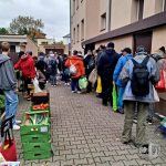 Ludzie czekają w kolejce przed budynkiem, aby otrzymać jedzenie ze skrzynek z warzywami umieszczonych na ziemi.