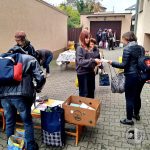 Ludzie przeglądający i wymieniający przedmioty na wyprzedaży podwórkowej, ze stołami i skrzyniami z towarami na utwardzonym terenie.