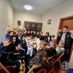 Grupa ludzi siedzących wokół stołu, uśmiechających się i podnoszących ręce w kierunku kamery.