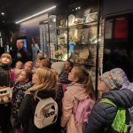 Grupa dzieci z plecakami ogląda eksponaty muzealne za szklanymi witrynami.