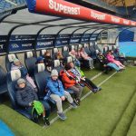 Dzieci siedzą na ławce drużyny piłkarskiej na stadionie, ubrane w kurtki i czapki, z dorosłym w pobliżu.