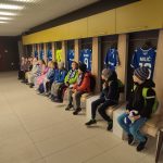 Grupa dzieci siedzi na ławkach w szatni, a za nimi wiszą niebieskie koszulki piłkarskie.