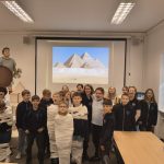 Grupa uczniów w klasie, niektórzy owinięci w biały papier, z piramidami wyświetlanymi na ekranie za nimi.