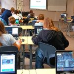 Uczniowie w klasie pracują na laptopach, stojąc przed ekranem wyświetlającym Code week.