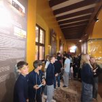 Grupa dzieci w szkolnych mundurkach stoi i przygląda się eksponatom w jasnym korytarzu muzeum o żółtych ścianach.