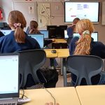 Uczniowie w klasie siedzą przy biurkach z laptopami, pracując nad projektami kodowania wyświetlanymi na ekranach.