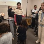 Uśmiechnięta dziewczyna owinięta białym materiałem jak mumia, otoczona przez kolegów z klasy.