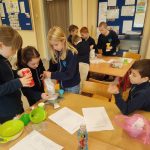 Dzieci w klasie pracują razem przy stołach, odmierzając składniki i czytając z wydrukowanych arkuszy.